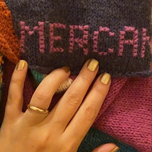 May include: A close-up of a hand wearing a gold ring, holding a multi-colored knitted blanket with the word "MERCI" embroidered in pink on a dark purple section.