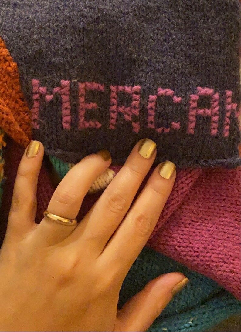 May include: A close-up of a hand wearing a gold ring, holding a multi-colored knitted blanket with the word "MERCI" embroidered in pink on a dark purple section.