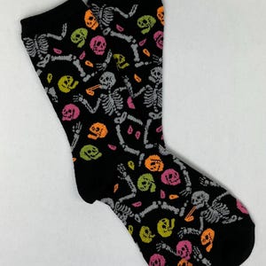 May include: A pair of black socks with a repeating pattern of gray skeletons and colorful skulls in orange, pink, and green. The socks are made of a soft material and are suitable for everyday wear.