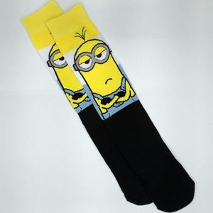 May include: A pair of knee-high socks featuring a yellow Minion character design. The socks have a yellow cuff, a white and blue background, and a black foot. The Minion has gray goggles and a grumpy expression.