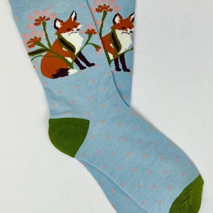 May include: A pair of light blue socks with a repeating pattern of small pink dots. Each sock features a fox illustration with orange fur, white chest, and black accents, perched among pink flowers. The heel and toe are green.