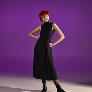Peut inclure: Robe noire sans manches avec un col montant et une fermeture asymétrique à boutons. La robe arrive à mi-mollet et est portée avec des bottines noires. Le fond est un dégradé de violet et de blanc.
