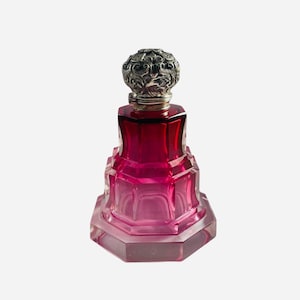 Può includere: Un flacone di profumo in vetro rosso rubino sfaccettato con base ottagonale a gradini e corpo a livelli. Il flacone è sormontato da un tappo rotondo in metallo color argento ornato da intricati motivi floreali e volute in rilievo.