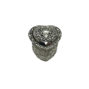 May include: A silver heart-shaped trinket box with ornate detailing on the lid. The box has a textured, faceted base. The lid features a detailed floral design. The box is likely made of metal.