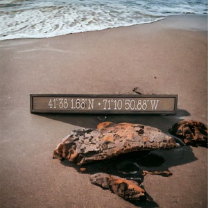 May include: A wooden sign with the coordinates "41°38'1.68"N - 71°10'50.88"W" written in white text. The sign is on a sandy beach with rocks and ocean waves in the background.