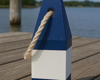 May include: A nautical-themed wooden buoy painted in navy blue and white, with a natural rope handle. The buoy is rectangular with a trapezoidal top. The background shows a wooden dock and a body of water.