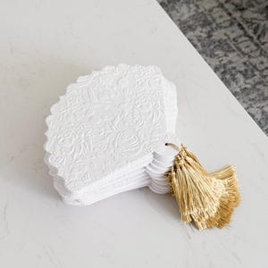 Könnte beinhalten: Ein Stapel weißer Karten mit wellenförmigen Kanten und geprägten Blumenmustern. Jede Karte hat ein kleines Loch und ist mit einer goldenen Quaste versehen. Die Karten liegen auf einer weißen Oberfläche.