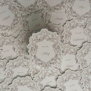 Könnte beinhalten: Eine Sammlung von cremefarbenen Platzkarten aus Papier mit einem schildförmigen Design und filigranen Rosen- und Schleifen-Linienzeichnungen in metallischem Roségold. Jede Karte zeigt das Wort WELCOME und einen individuellen Namen.