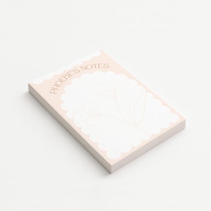 May include: A rectangular notepad with a pale pink border and a scalloped edge. The notepad has the text "PHOEBE'S NOTES" in a light brown colour. A floral design is visible on the white paper.