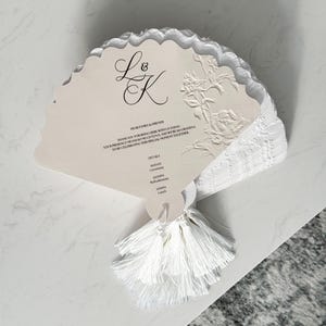 May include: A pile of ivory wedding fan invitations with a scalloped edge. The fans have embossed floral designs, the initials "L & K", and event details. A white tassel hangs from the bottom of the stack.