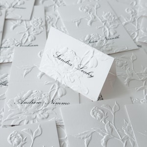 May include: A collection of white folded place cards featuring an embossed floral design of peonies and leaves. Each card displays a unique name printed in black script, such as Sandra Leahy, Andrew Nimmo, and Sean Nasseth, on the textured surface.