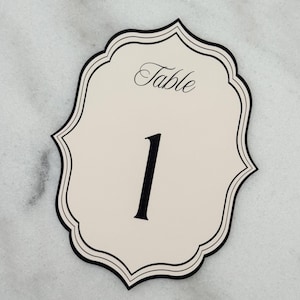 May include: A cream-coloured table number card with a decorative scalloped edge and black border. The card features the word Table in a black script font at the top and the large black numeral 1 centred below it on a smooth, flat surface.