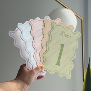 Puede incluir: Un juego de cinco tarjetas de número de mesa con bordes ondulados en colores pastel que incluyen verde, amarillo, rosa, azul y morado. Cada tarjeta presenta la palabra Table en letra cursiva sobre un número grande, con el 1 en la tarjeta verde.