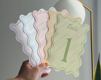 May include: A set of five wavy-edged table number cards in pastel colors including green, yellow, pink, blue, and purple. Each card features the word Table printed in a script font above a large numeral, with the green card showing the number 1.