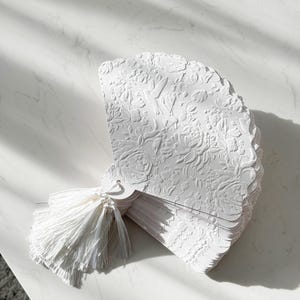 May include: A stack of white paper fans featuring an intricate embossed floral and bird pattern across the surface. Each fan has a scalloped edge and is bound together at the base with a white decorative tassel made of thin, soft fabric threads.