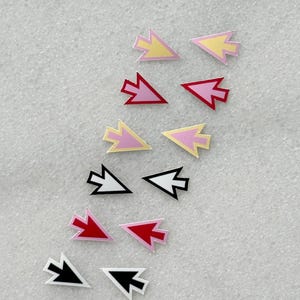 May include: A selection of small, arrow-shaped embellishments in various colours. The arrows feature pink, yellow, red, black, and white outlines and fills. The background is a light, textured surface.