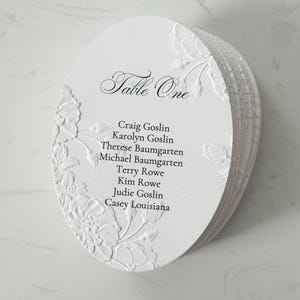 May include: A stack of oval white cardstock table seating cards featuring embossed floral patterns along the edges. The top card displays the text Table One followed by a list of names including Craig Goslin, Karolyn Goslin, Therese Baumgarten, Michael Baumgarten, Terry Rowe, Kim Rowe, Judie Goslin, and Casey Louisiana.