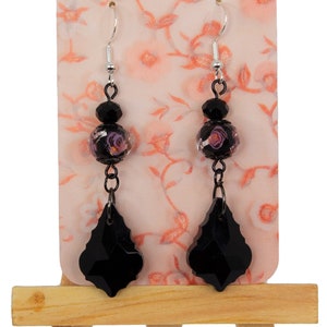 May include: Black dangle earrings with a teardrop-shaped crystal and a round bead with a floral pattern. The earrings are displayed on a small wooden easel.