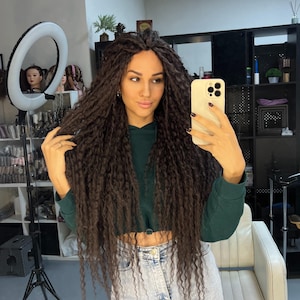 Puede incluir: Una mujer con cabello largo y rizado de color marrón oscuro, que lleva una sudadera con capucha corta verde oscuro y vaqueros azul claro, se hace un selfie en un salón bien iluminado. Un aro de luz y espejos son visibles en el fondo, junto con estantes de extensiones de cabello.