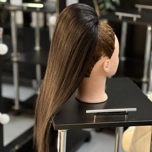 May include: A mannequin head with brown hair styled in a high ponytail. The hair is long and straight, with a dark brown color at the roots that fades into a lighter brown. The mannequin is on a black table.