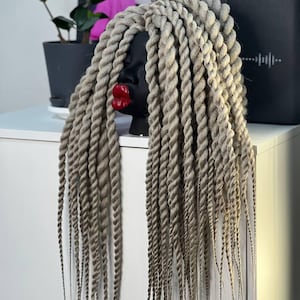 Può includere: Una testa di manichino nera con labbra rosse e lunghe trecce attorcigliate grigio chiaro. Le trecce scendono a cascata, mostrando la consistenza e lo stile dei capelli. La testa è posizionata su una superficie bianca.