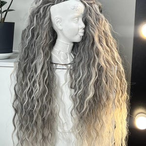 Pode incluir: Um busto de cerâmica branca com uma longa peruca ondulada em tons de cinza e loiro. A peruca é estilizada com a parte superior raspada e cachos longos e fluidos. O busto tem detalhes faciais e marcas decorativas na cabeça.