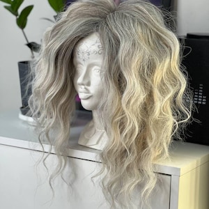 May include: A wavy, light blonde wig with gray roots is displayed on a white mannequin head. The wig has long, flowing locks with a natural-looking texture. The mannequin is on a white surface.