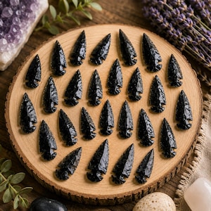 Puede incluir: Una colección de veinticinco puntas de flecha de obsidiana negra dispuestas sobre una rodaja de madera circular. Cada punta presenta un extremo afilado y una base con muescas, con una textura brillante y escamada.