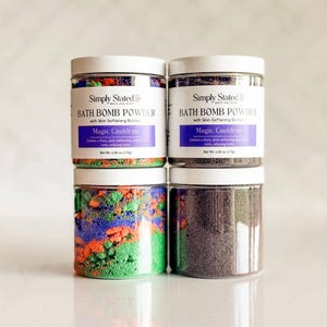 Puede incluir: Dos frascos de plástico transparente con polvo para bombas de baño Simply Stated, aroma Magic Cauldron. El frasco izquierdo muestra capas de polvo verde, naranja y morado. El frasco derecho contiene polvo morado oscuro. Peso neto 170g.