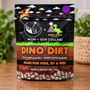 May include: A black pouch of Dino Dirt fizzy bath powder with a Dino Keys surprise. The packaging features a cartoon dinosaur, a volcano, and a crescent moon. Text includes "MOM + SON COLLAB!", "DINO DIRT", "Fizzy bath powder + DINO KEYS surprise", and "142g".