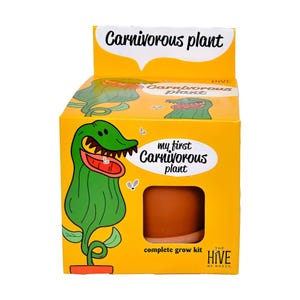 以下が含まれることがあります： 「My First Carnivorous Plant」の完全栽培キットが入った黄色い段ボール箱。箱には、大きな口と鋭い歯を持つ緑色の漫画風の植物、小さなハエ、そして中のテラコッタ色のプラスチック製ポットが見える切り抜き窓が描かれています。