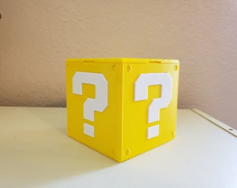 May include: A yellow cube with a question mark on each side. The cube is made of plastic and has a smooth surface.