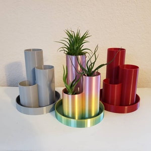 May include: Three 3D printed desk organisers, each with a different colour scheme. The first is grey, the second is a rainbow gradient, and the third is red. Each organiser has multiple compartments for pens, pencils, and other office supplies. The organisers are on a white surface.