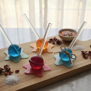 May include: A wooden cutting board displays colorful lollipops with clear sticks. The lollipops are blue, orange, and red, resting on star-shaped holders. A small bowl of sprinkles and decorative elements complete the scene.