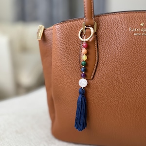 May include: A brown leather handbag with a gold-tone metal logo reading kate spade NEW YORK. Attached to the handle is a decorative charm featuring a gold ring, a vertical row of multi-colored gemstone beads, and a dark blue fabric tassel.