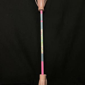 May include: A handmade pink and floral patterned pencil with a decorative tassel at each end. The tassels are made of layered paper. The pencil is pink and clear with a floral pattern. The background is black.