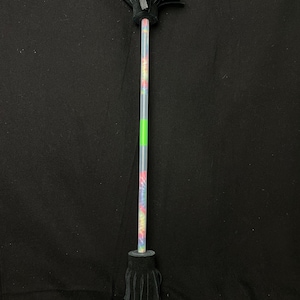 May include: A tie-dye patterned wand with black leather fringe at both ends. The wand has a green band and a black handle. The background is a solid black color.