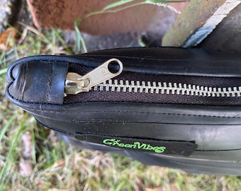 May include: Black bag with a silver zipper and the text "Green Vibes" in green. The bag is made of a dark material and is partially open, revealing the zipper teeth.