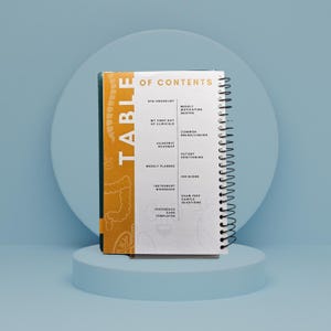 May include: A spiral-bound notebook with a white cover and black spiral binding. The cover features the word "TABLE" in gold lettering and a decorative pattern. The notebook's contents include checklists, weekly planners, and exam prep questions.