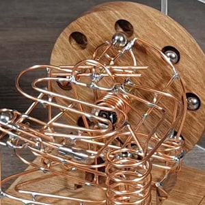May include: A wooden and copper kinetic sculpture with a complex wire track for steel balls. The sculpture is mounted on a rectangular wooden base. A laptop and a clear acrylic cube are in the background.