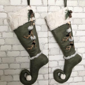 May include: Two green elf-style Christmas stockings with white faux fur cuffs and brown antler accents. The stockings are decorated with pine cones, berries, and white pom-poms.