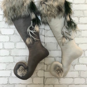 May include: Two Christmas stockings, one grey and one off-white, with faux fur trim and decorative silver antlers. Each stocking has a curled toe with a pom-pom and a green hanging loop.