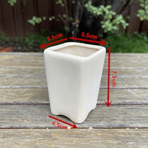 May include: A white ceramic planter with a square shape and rounded edges. The planter measures 5.5 cm wide, 7.5 cm tall, and 4.5 cm deep.
