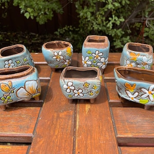 May include: Six small, blue ceramic flower pots with white and yellow floral designs. Each pot has four small feet and a brown interior.