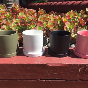 May include: Four ceramic plant pots with matching saucers in different colours: olive green, white, black, and pink. The pots are arranged in a row on a red wooden surface.