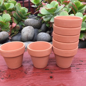 May include: A stack of eight terracotta flower pots, with two additional pots in the foreground. The pots are all the same size and shape, and they are arranged in a staggered pattern. The pots are sitting on a wooden surface.