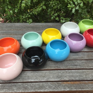 May include: Ten round ceramic planters in a variety of colours, including pink, orange, black, blue, green, yellow, white, and purple. The planters are arranged in a row on a wooden surface.