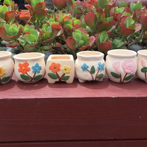 May include: Six small ceramic flower pots with different floral designs. The pots are white with colourful flowers and leaves.