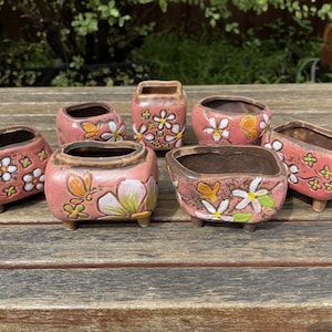 May include: Seven small, pink ceramic planters with a distressed finish. Each planter features white flowers, green leaves, and orange butterflies. The planters have a brown interior and sit on small, brown feet.