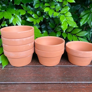 May include: A set of terracotta pots in various sizes. The pots are unglazed and have a natural brown colour. The pots are stacked on top of each other, with the smallest pot on top. The pots are sitting on a wooden surface.